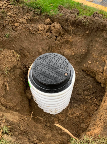 A black circular access lid sits atop a white corrugated plastic pipe installed in an open dirt excavation.