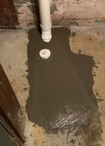 A white PVC pipe and cleanout cap embedded in fresh, wet gray concrete against an unfinished basement wall.