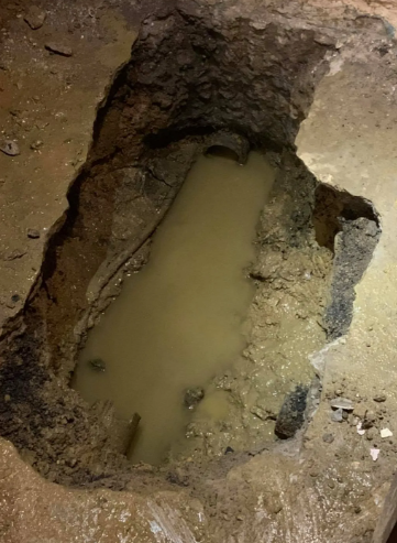 A deep, narrow excavation pit in the ground filled with murky, brown water, revealing a section of buried piping.
