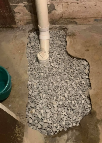 A white PVC pipe enters a concrete basement floor through a patch filled with grey gravel.