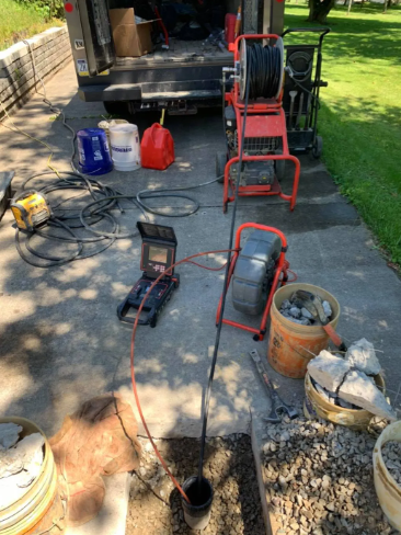 Sewer inspection equipment, including cameras and reels, sits on a driveway next to an open pipe in the ground.