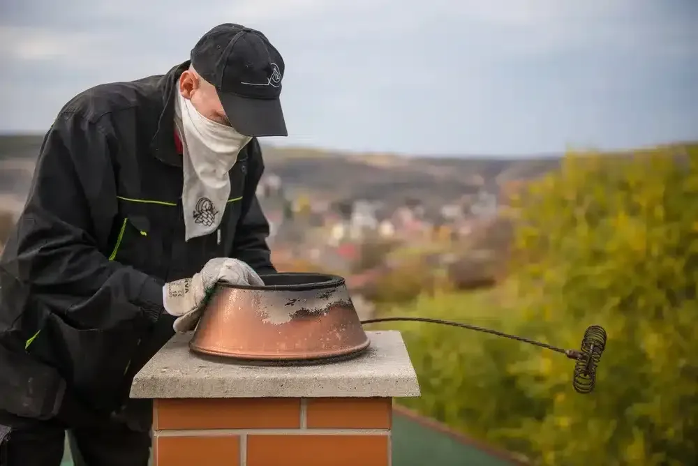 Person inspecting a rooftop chimney cap with a tool, outdoors with a blurred town in the background