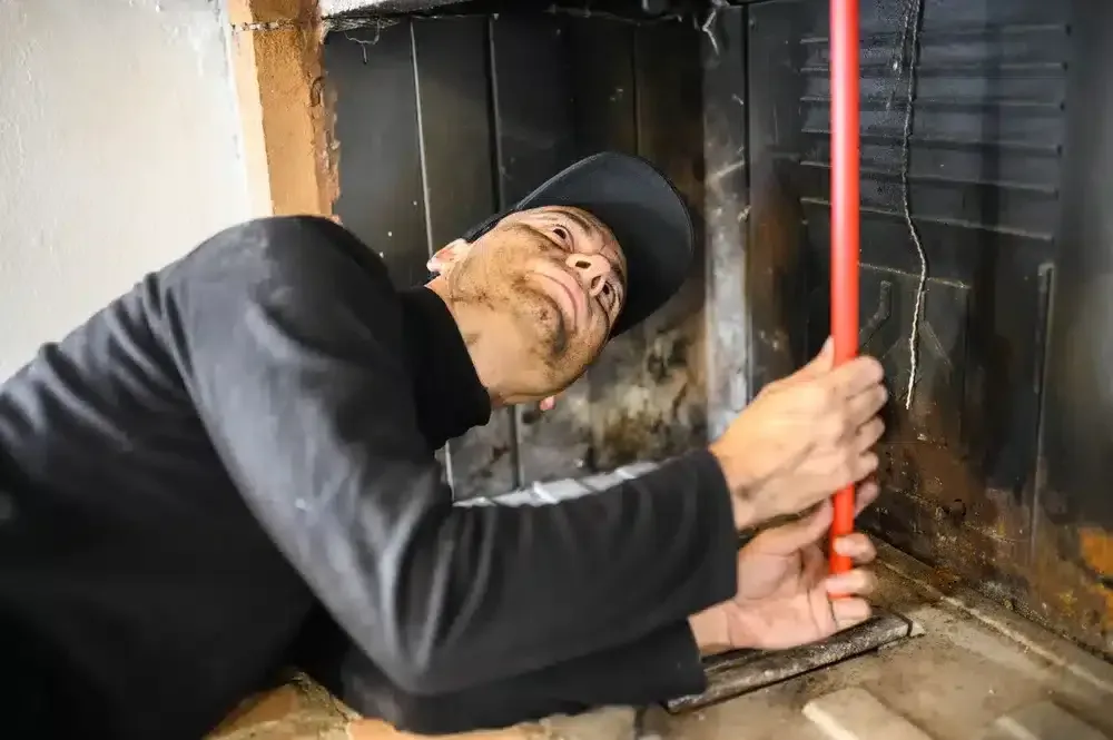 Worker in a black cap and coveralls reaching into a fireplace with a red rod