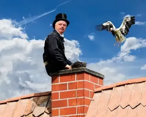 Man sitting on a brick chimney on a rooftop, with a bird flying nearby under a blue sky.