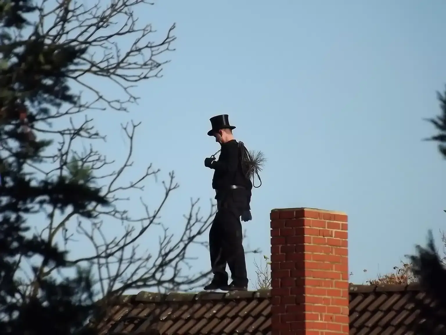 Person in a black outfit and top hat standing on a rooftop beside a brick chimney against a blue sky