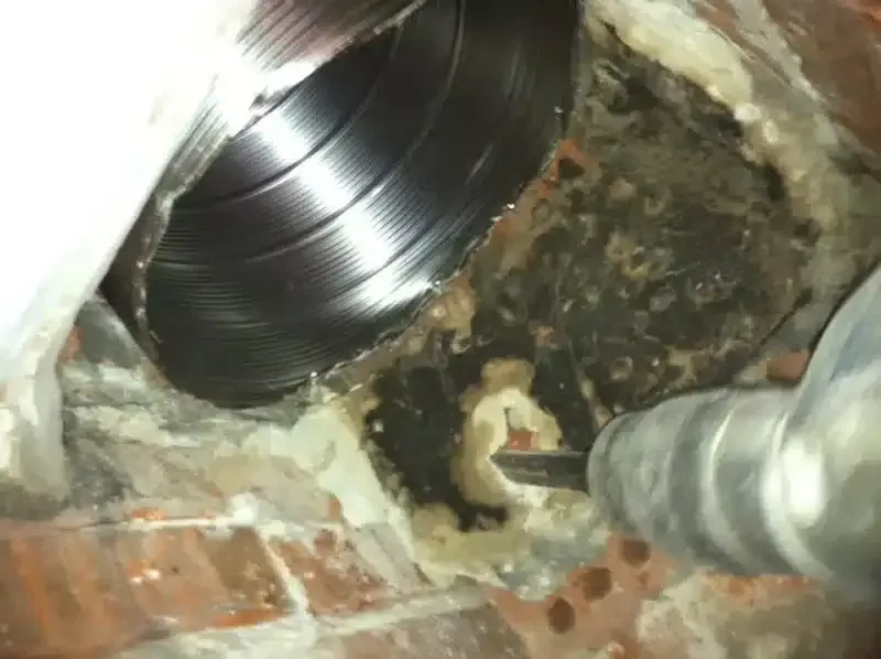 Inside a damaged wall opening, a shiny metal duct enters a dark, moldy cavity with pipes visible.