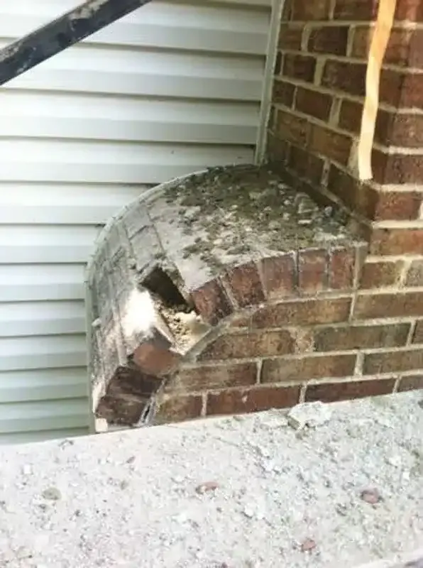 Close-up of a damaged brick arch ledge with a hole, beside white siding and a brick wall.