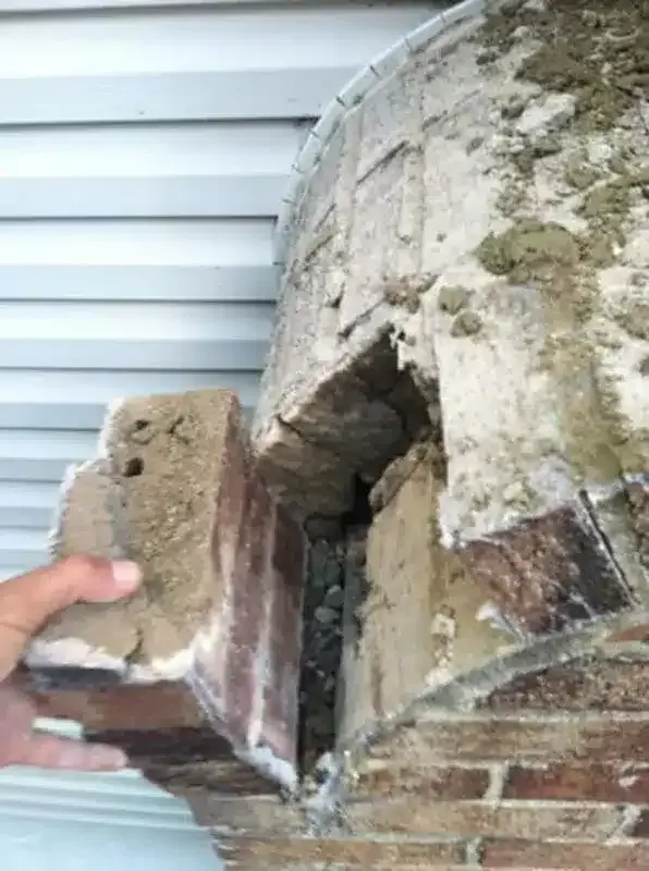 Hand holding a broken brick from a cracked, weathered chimney or wall.