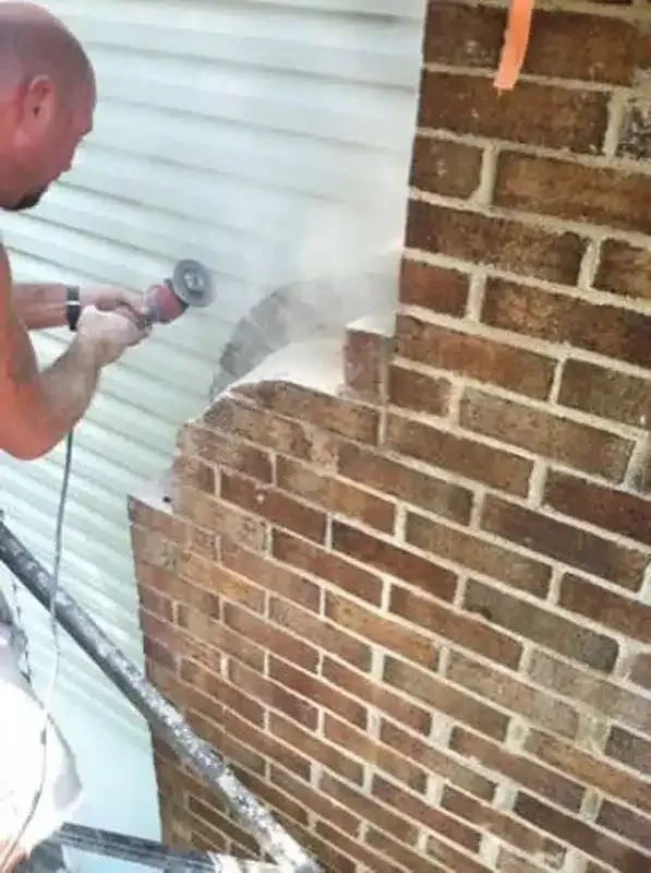 Man using a power grinder to cut into a brick wall beside white siding outdoors