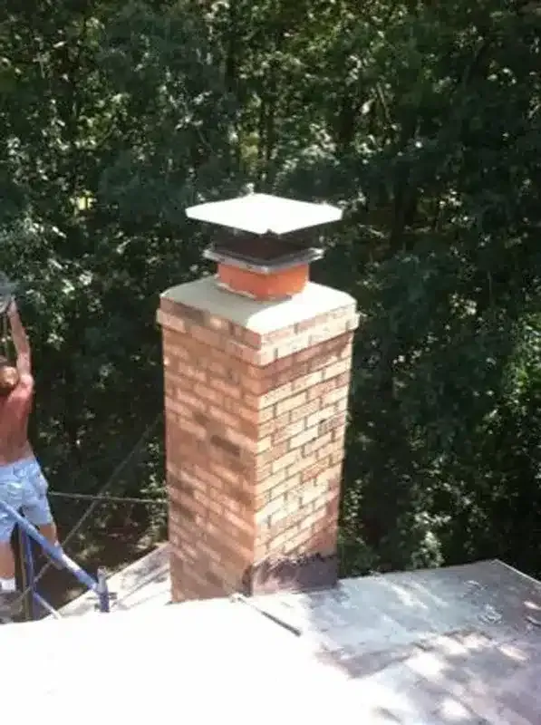 Brick chimney on a roof with a white cap, set against trees; a worker is partially visible at left.