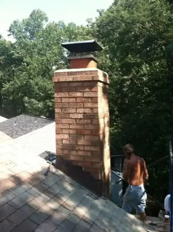 Brick chimney with metal cap on a shingled roof, with trees in the background and a worker nearby