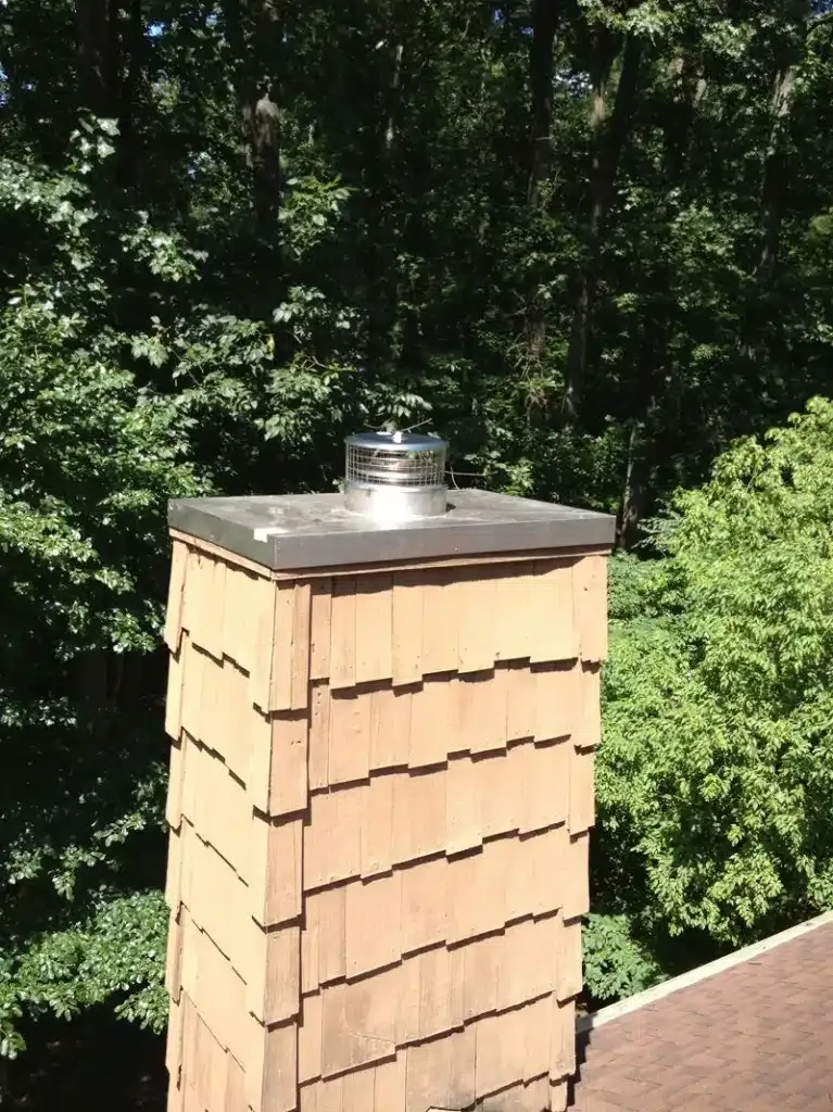 Tall beige chimney with metal cap on a roof, set against a leafy green background