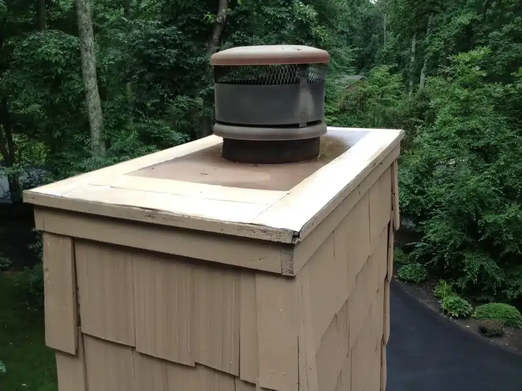 Tan vent on a tan shingled roof, surrounded by dense green trees.