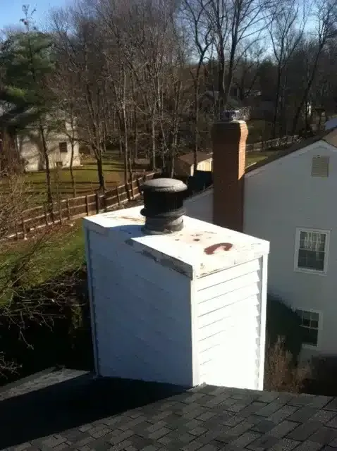 Rooftop chimney on a white house beside a tree-lined yard on a sunny day