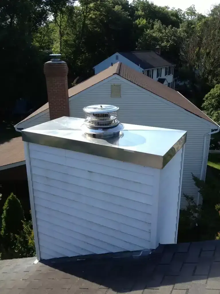 Rooftop vent on a small white dormer with a metal cap and brick chimney, backed by trees and a house.