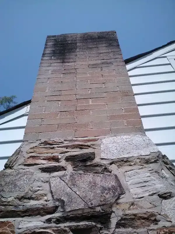Tall brick chimney with stone base against a blue sky and white-sided building