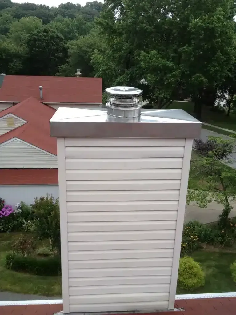 White-sided rooftop chimney vent with metal cap, set against red roofs and green trees.