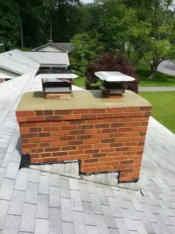 Brick chimney with two metal caps on a shingled roof, surrounded by green trees.