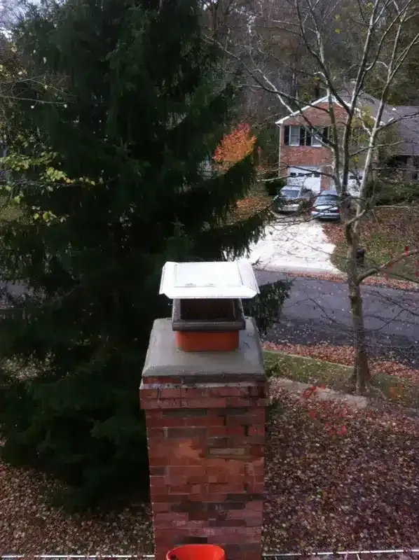 Brick chimney with white cap in a yard, with a tall evergreen and a house in the background.