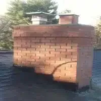 Brick chimney with a large curved crack near the base on a roof.