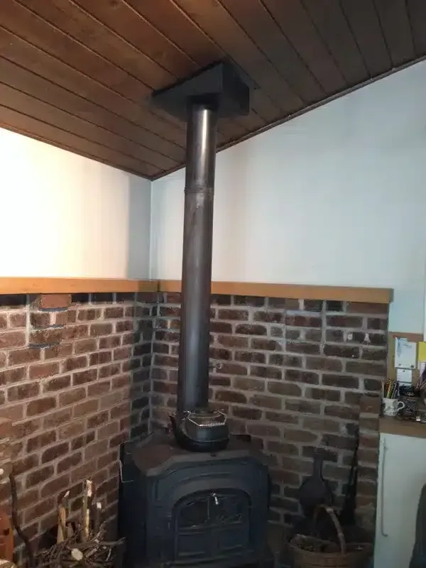 Brick corner with black wood stove, tall chimney pipe, and wood-paneled ceiling