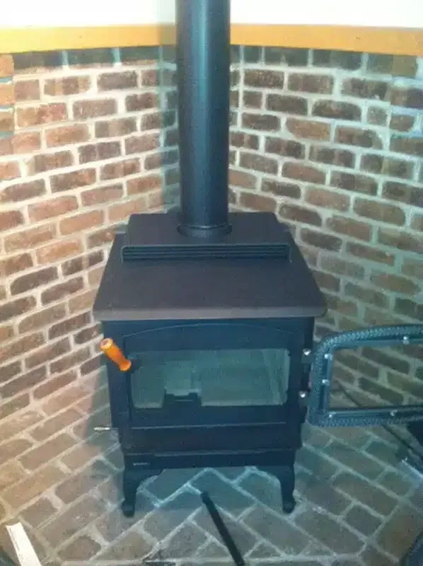 Black wood stove with chimney pipe in a brick corner room, with a hose on the right.