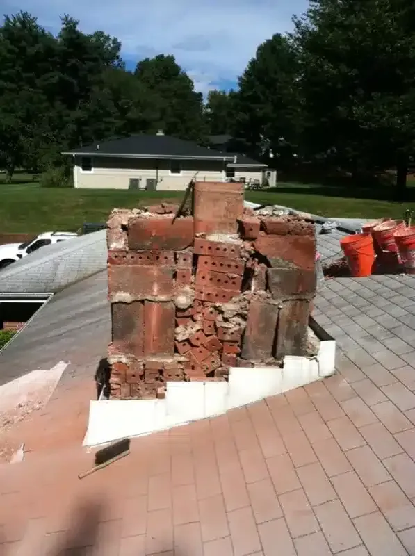 Crumbling brick chimney on a rooftop with white support base and orange debris bins nearby