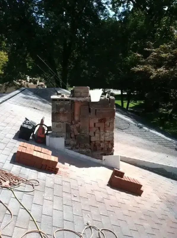 Partially built brick chimney on a sloped roof with loose bricks and tools nearby