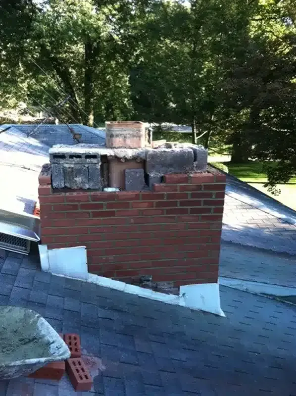 Brick chimney on a shingled roof with exposed top and a tree-lined yard in the background