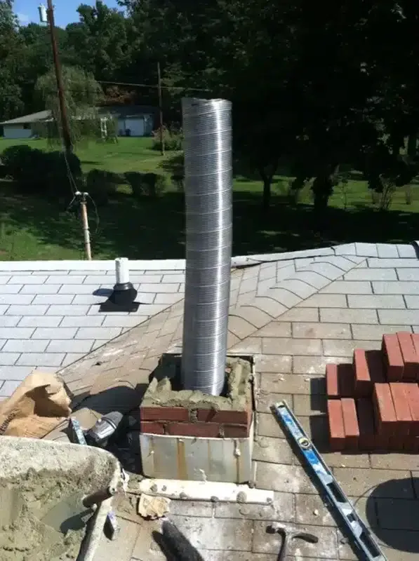 Silver chimney pipe protruding from a rooftop surrounded by shingles and bricks, with trees in the background