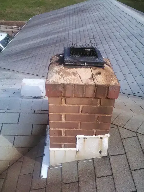 Brick chimney on a shingled roof with a black vent cap and a white utility box attached to the side