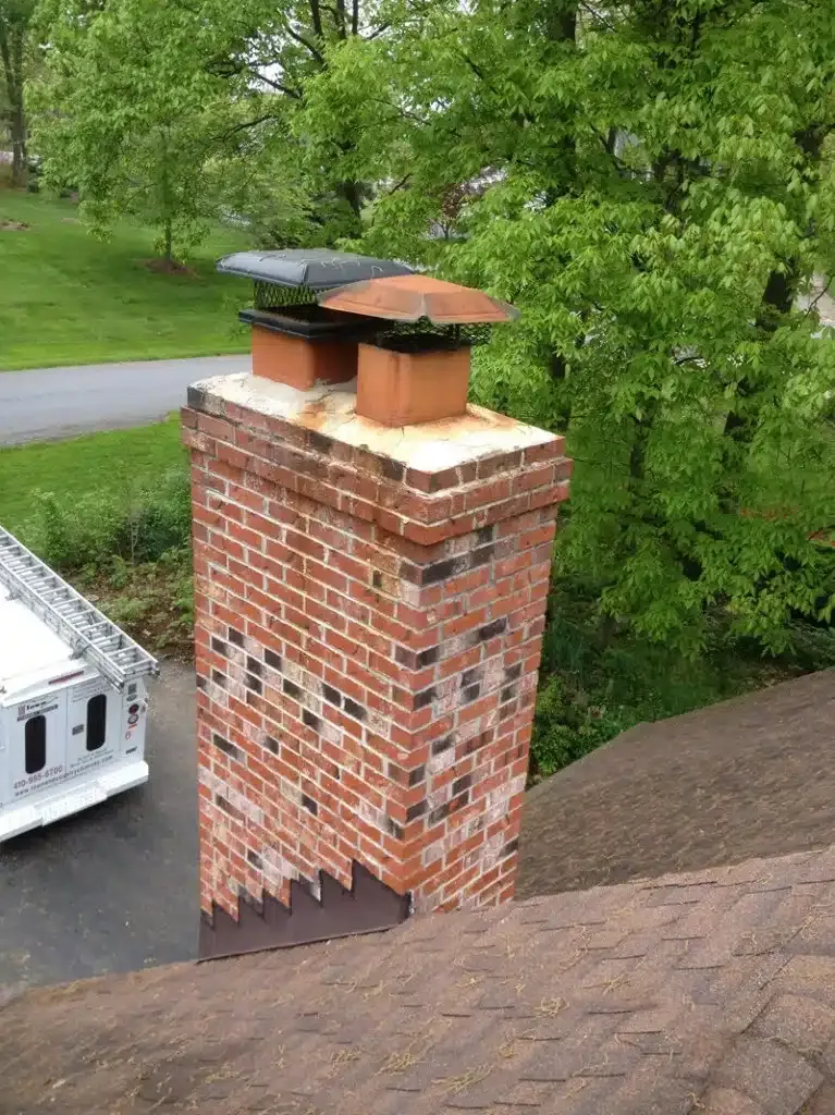 Brick chimney on a shingled roof beside a wooded road.
