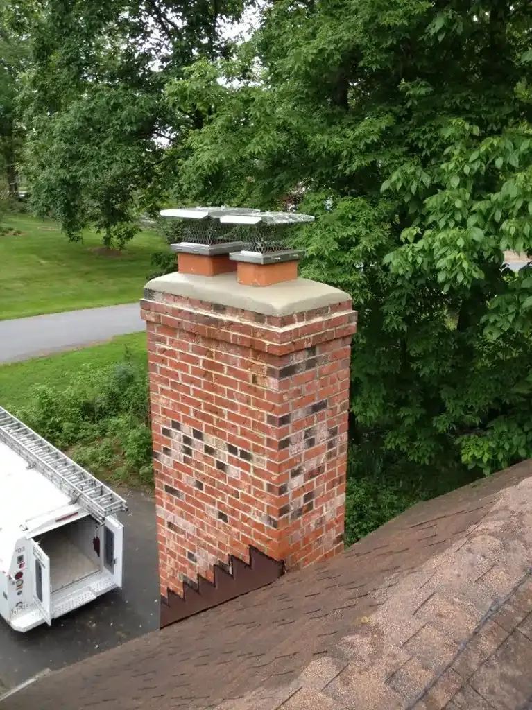 Brick chimney on a shingled roof beside a driveway, with trees in the background.