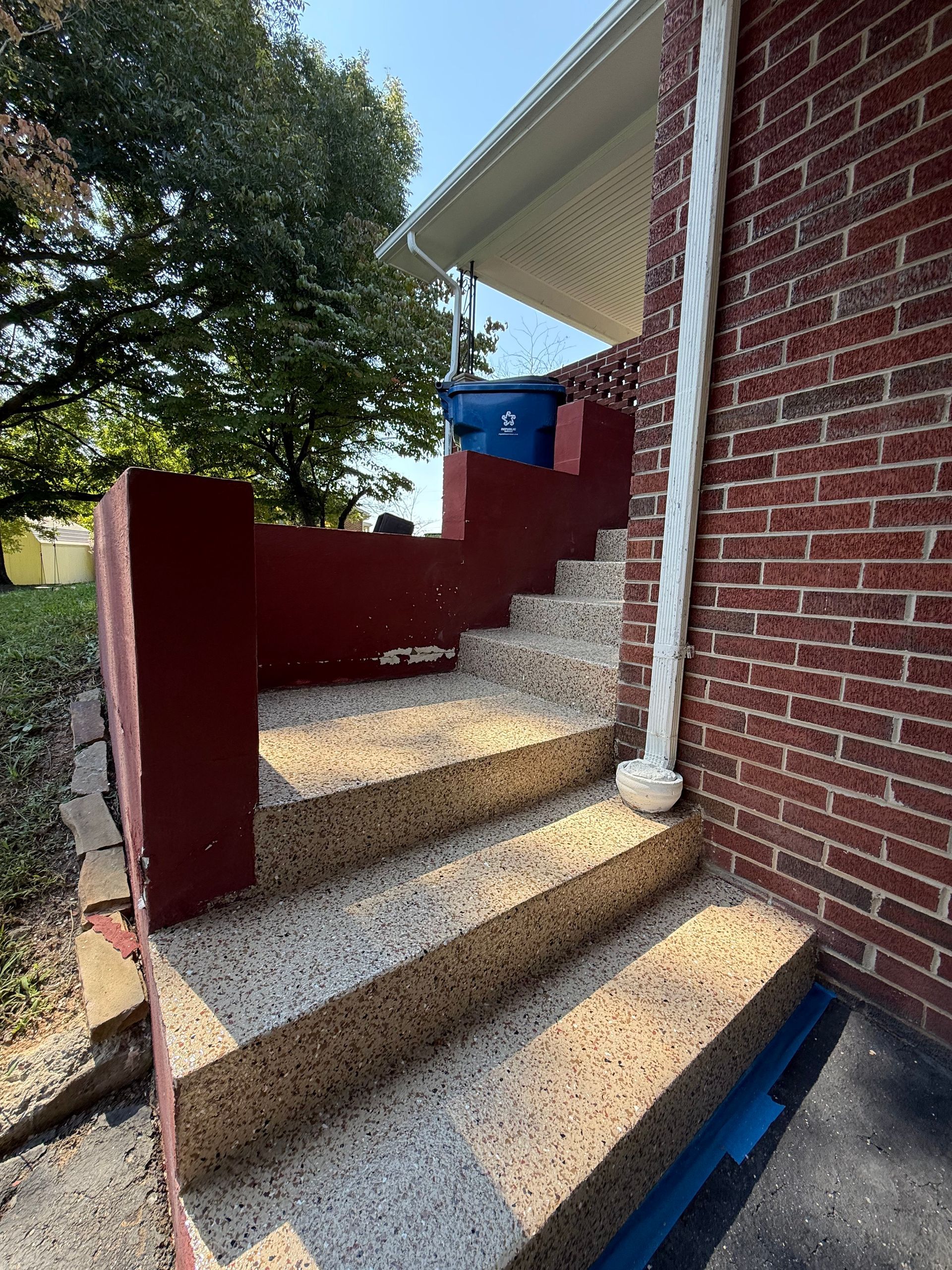 Brick house steps with red railings leading to a porch, with a blue bin at the top.
