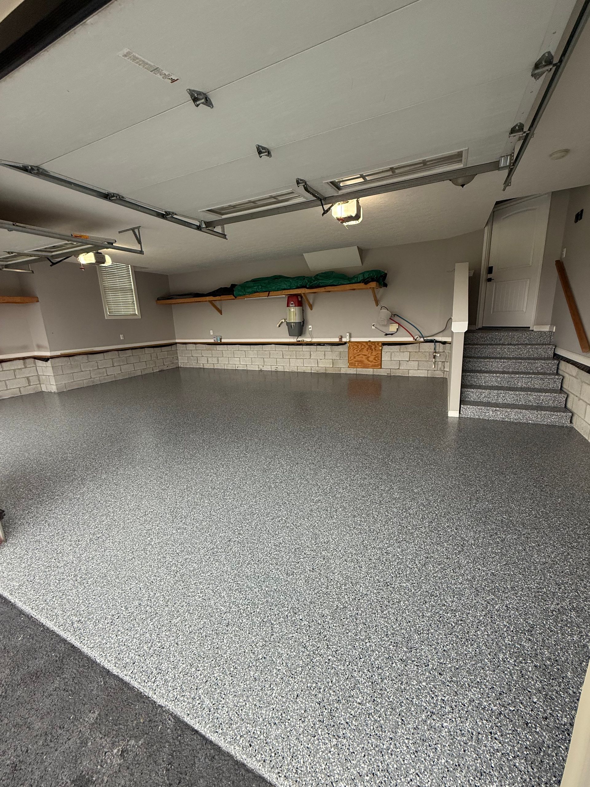 Empty garage with gray speckled floor, stairs on the right, and shelves along the back wall