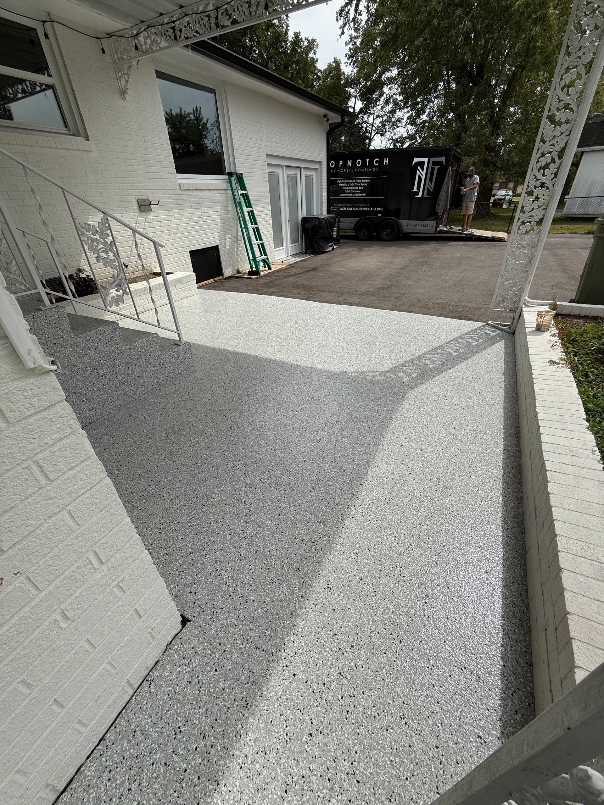 Outdoor gravel and concrete patio beside a white house, with a ladder and black truck in the background.
