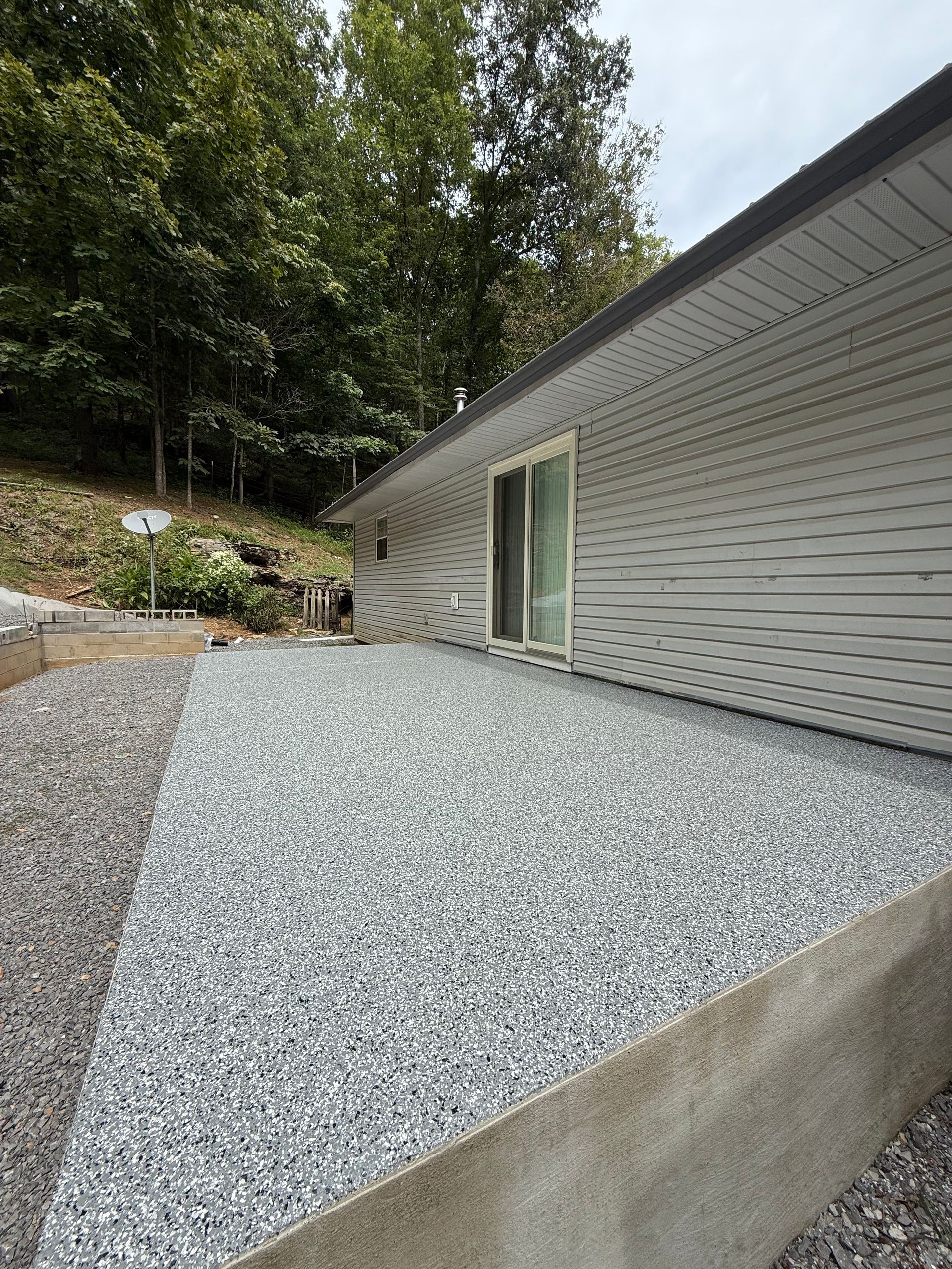 Gravel-covered concrete patio beside a light gray house with a sliding glass door and wooded hillside.