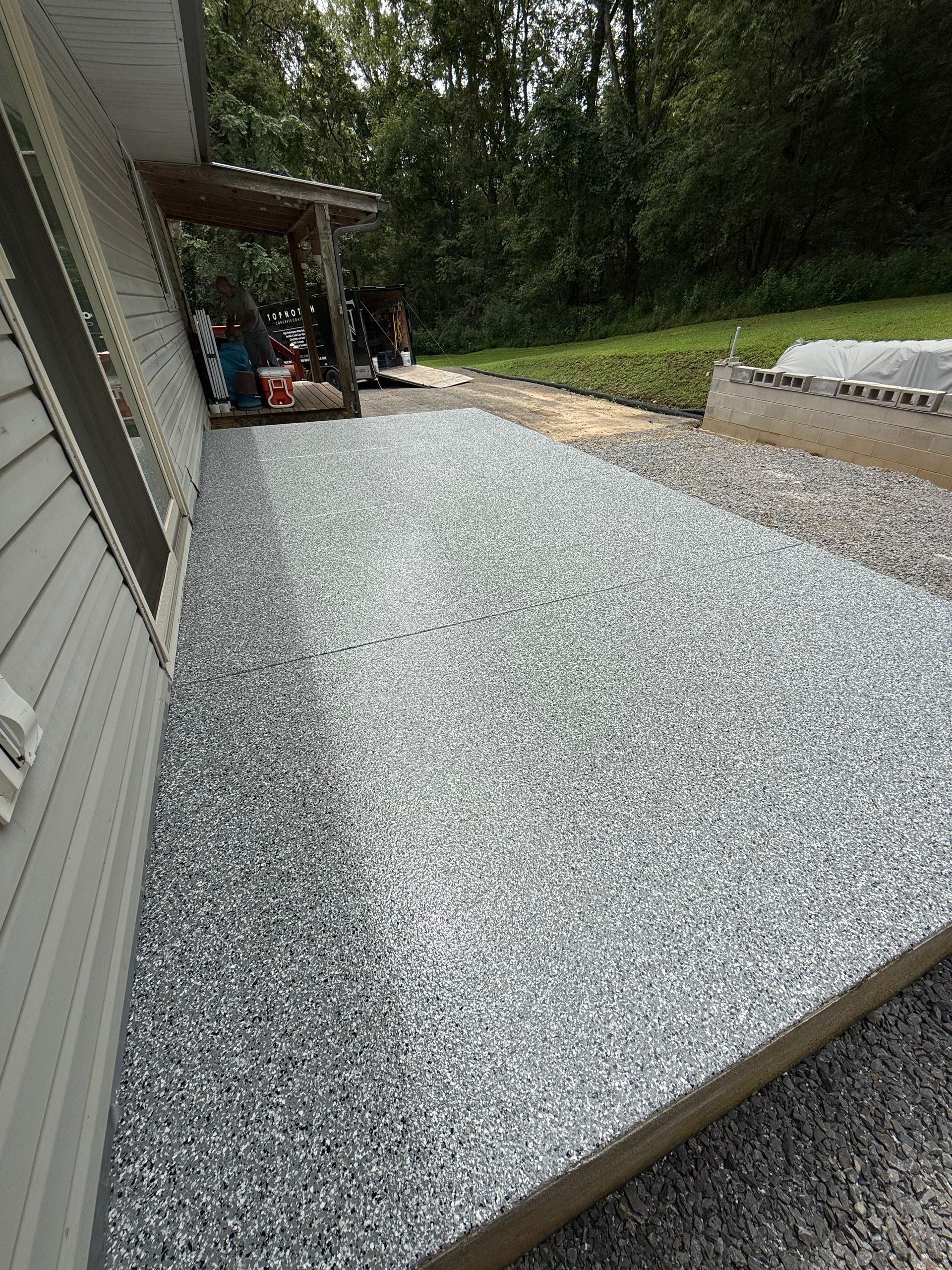 Gravel-covered sloped ramp beside a house, with a covered porch and trees in the background.