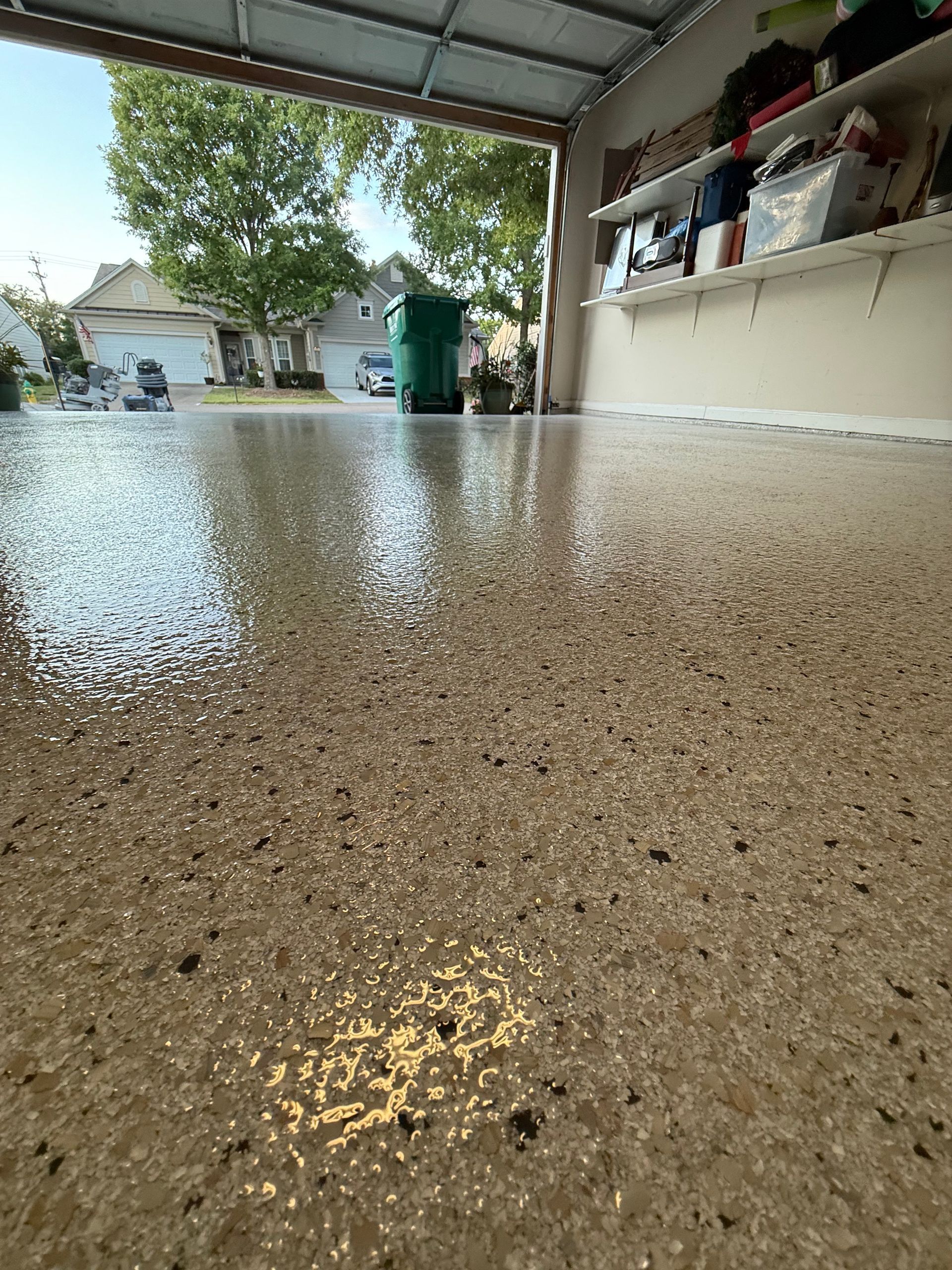 Low-angle view of a speckled garage floor reflecting light, with shelves, a green trash bin, and a yard outside