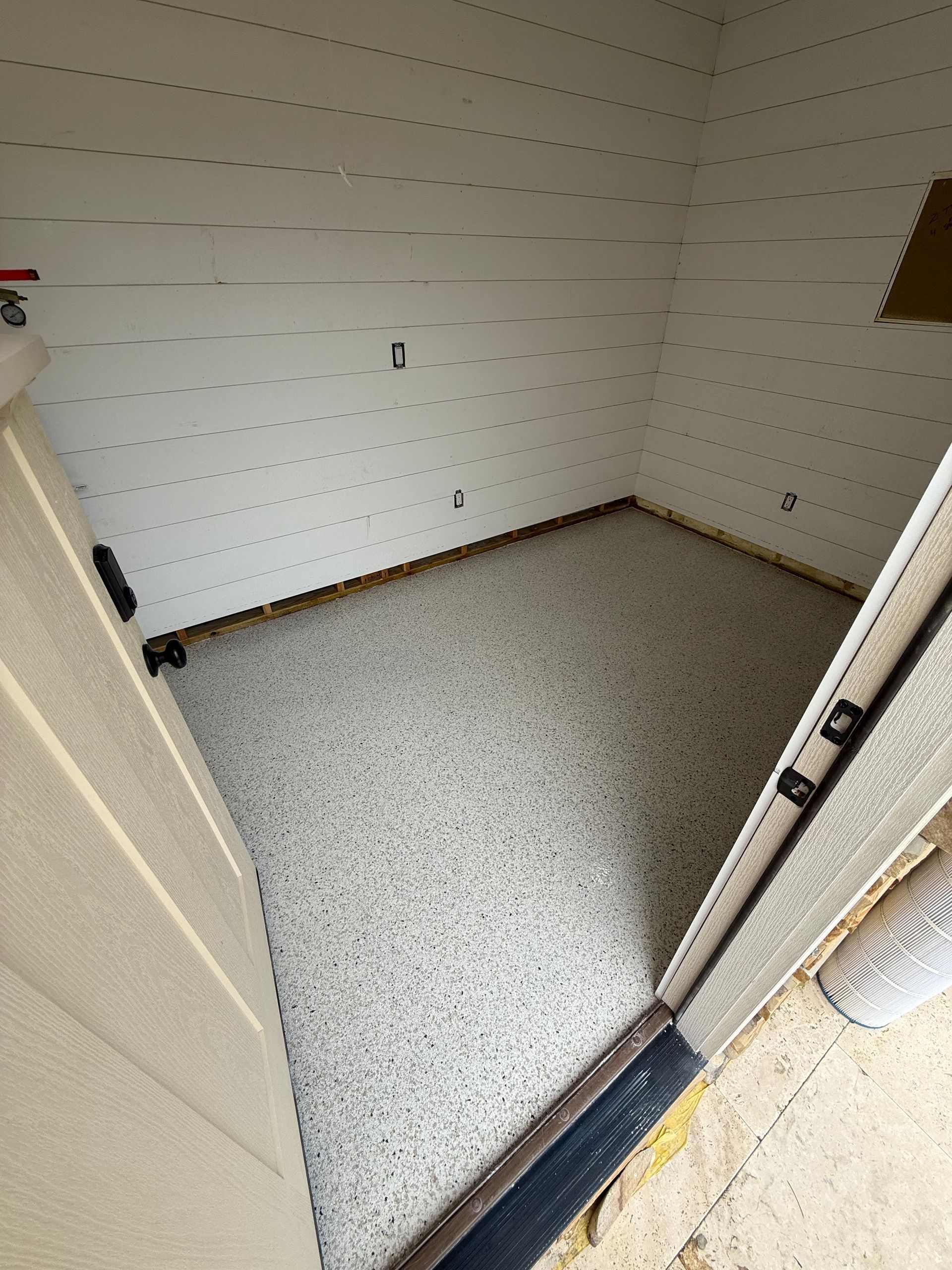 Empty small storage closet with white pegboard walls and speckled floor, viewed through an open door.