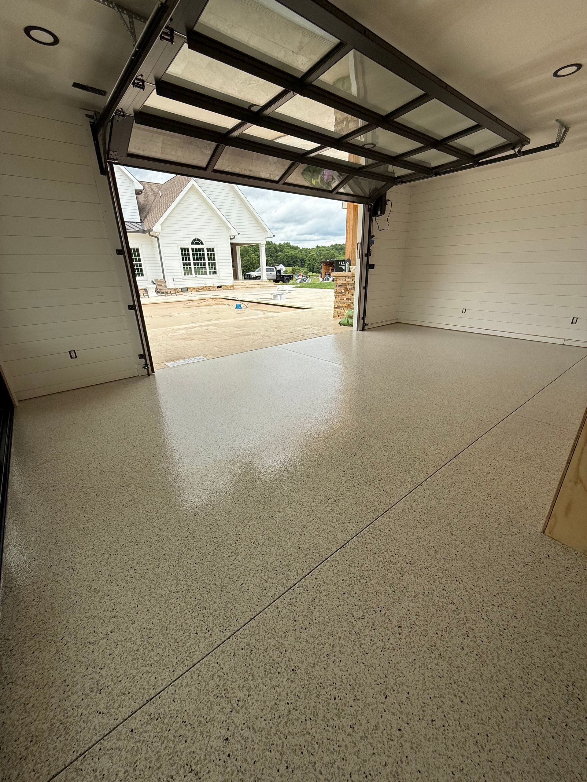 Empty garage with speckled floor and open door showing a house and driveway outside