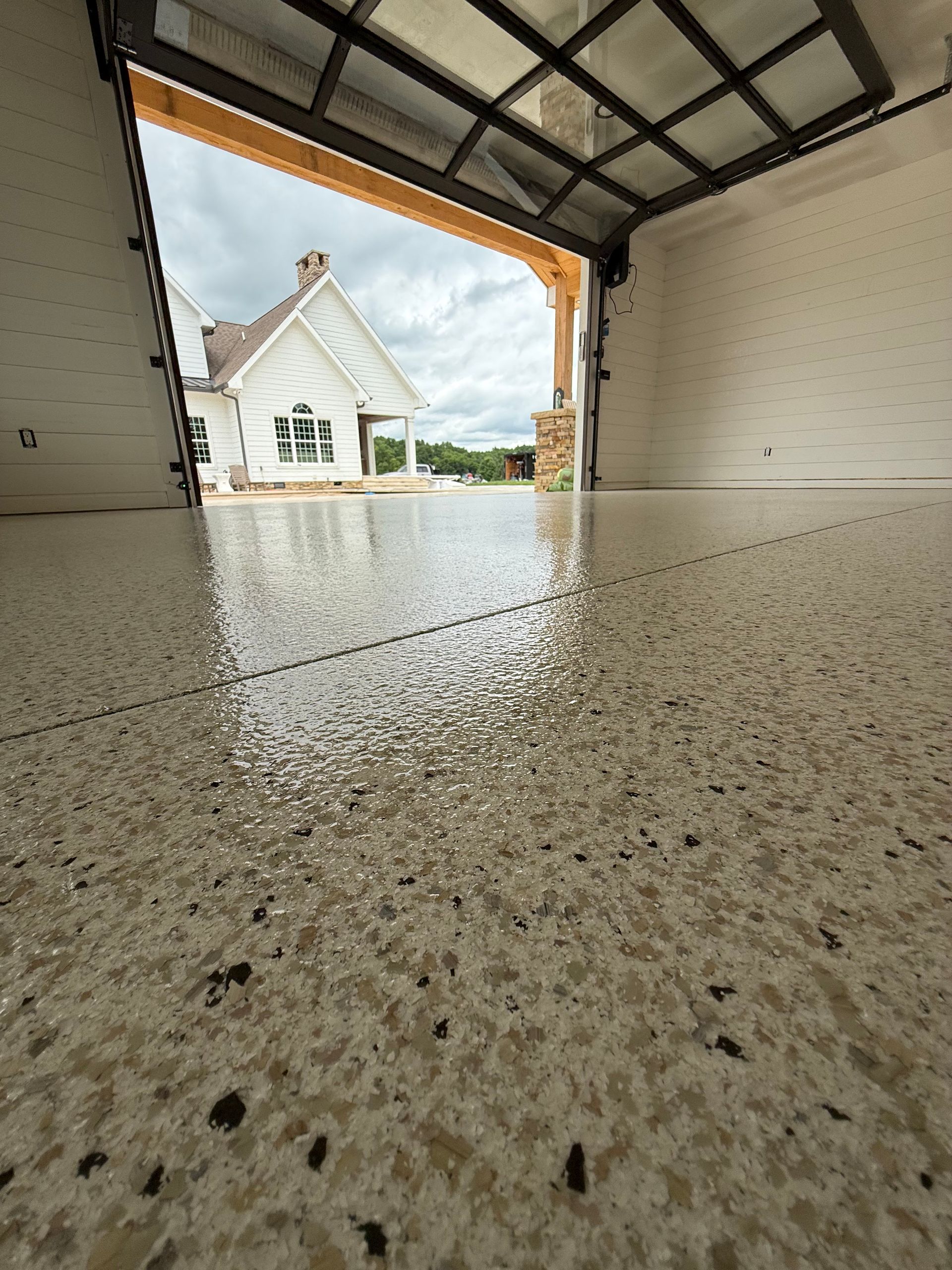 Empty garage interior with speckled floor, open door, and a house visible outside.