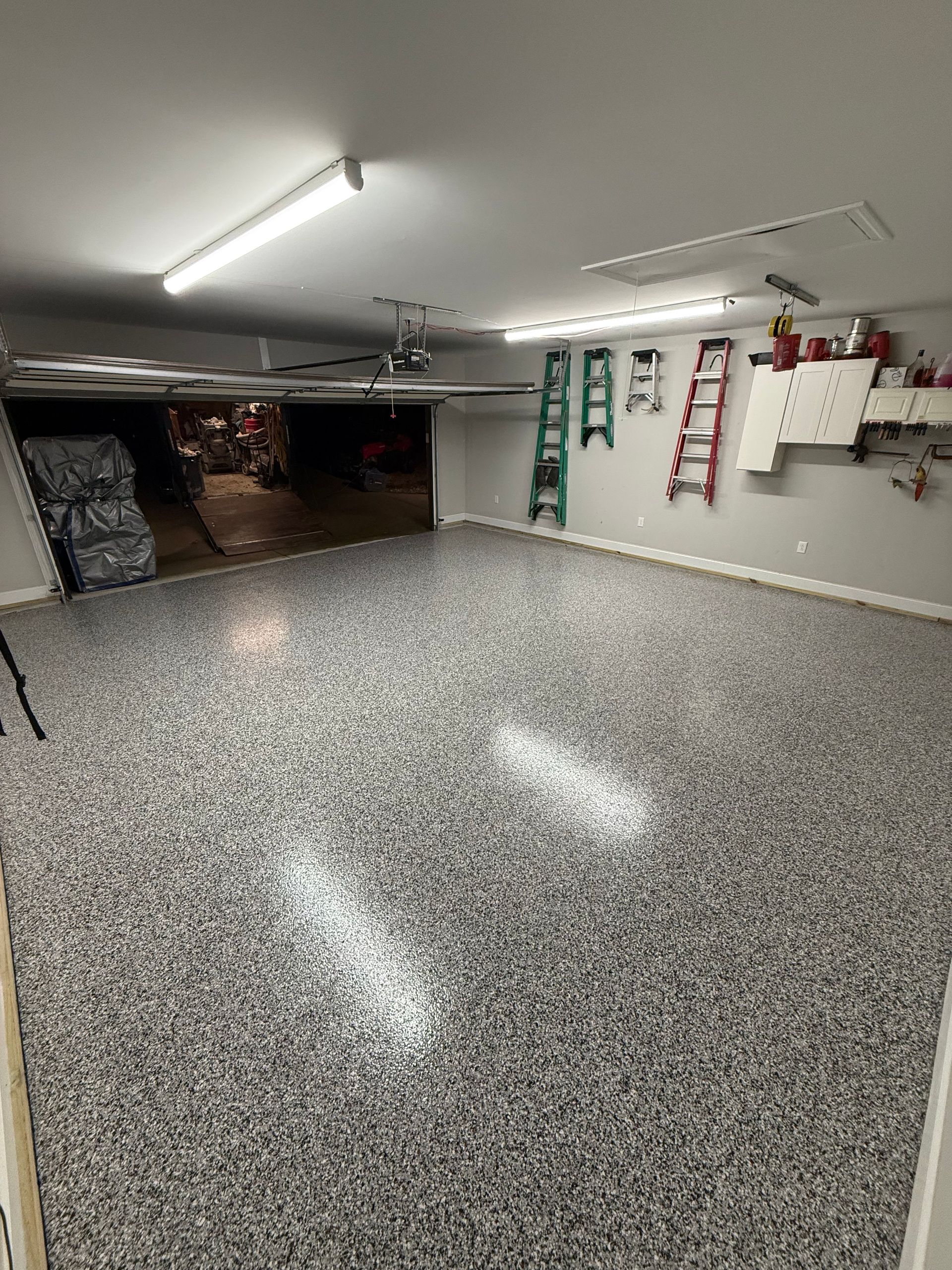 Garage with speckled epoxy floor, overhead lights, wall-mounted ladders, and shelves along the back wall.