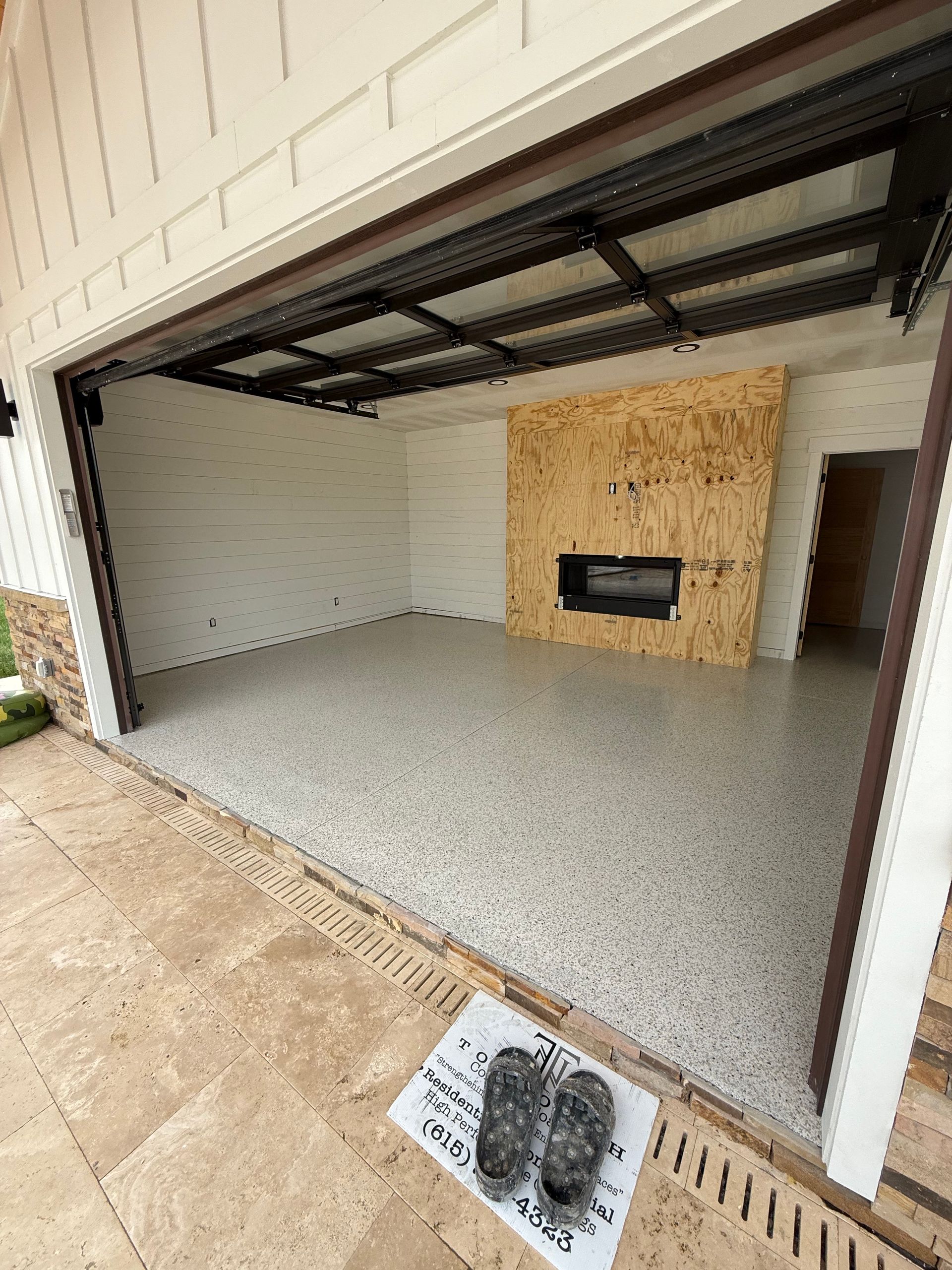Open garage with gray epoxy floor, white brick walls, and a plywood-covered fireplace on the back wall