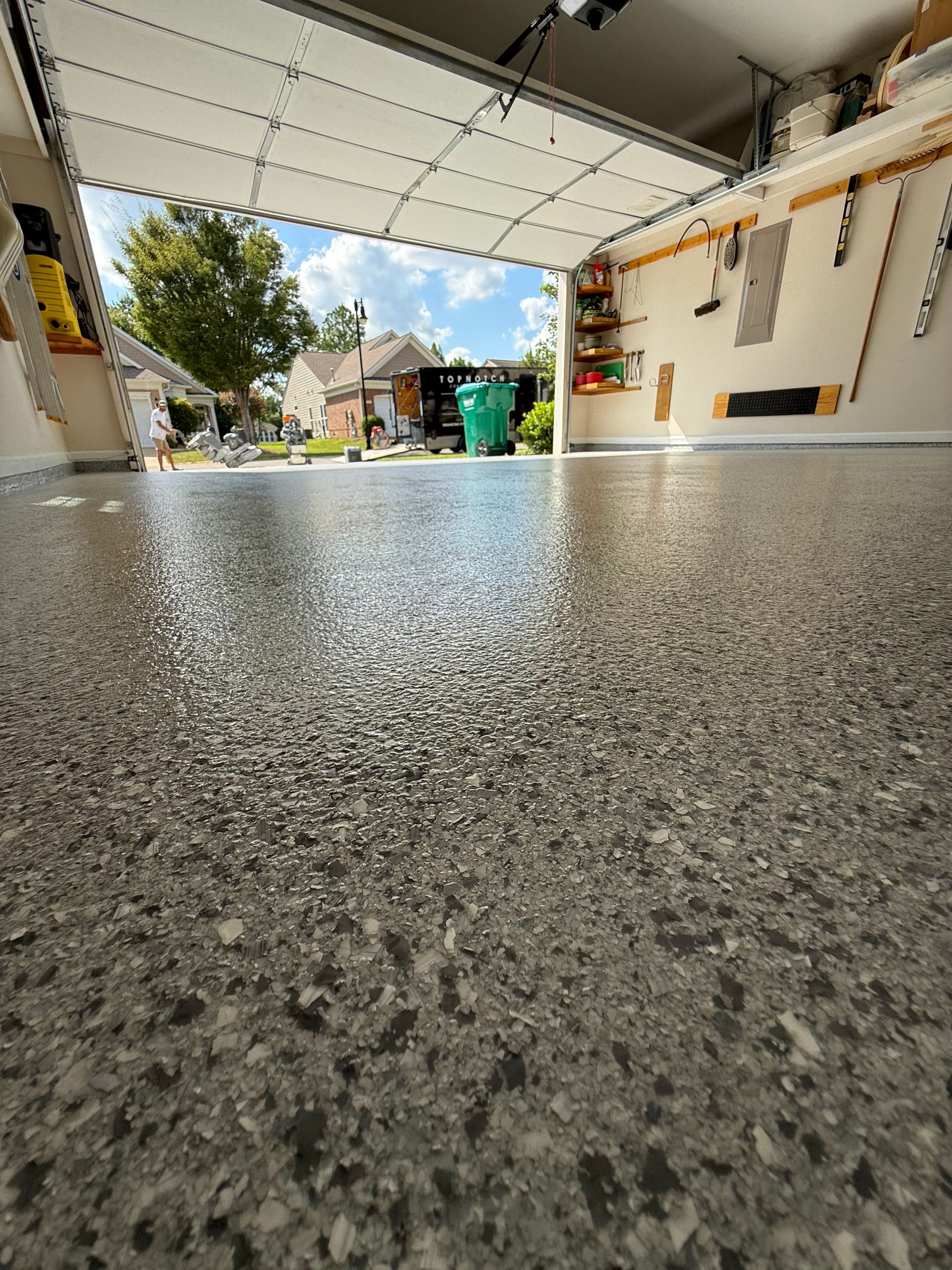 Low-angle view from a garage, showing a speckled floor and an open driveway with trees and houses beyond.
