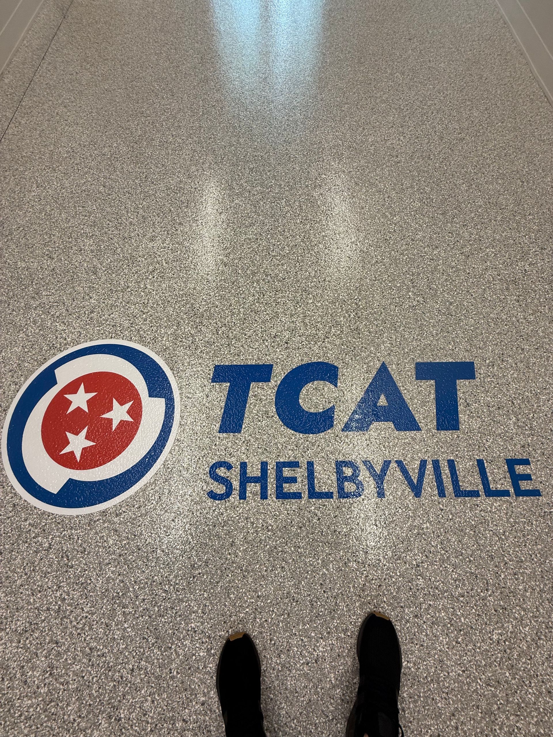 TCAT Shelbyville logo on a speckled floor, viewed from above with shoes visible at the bottom