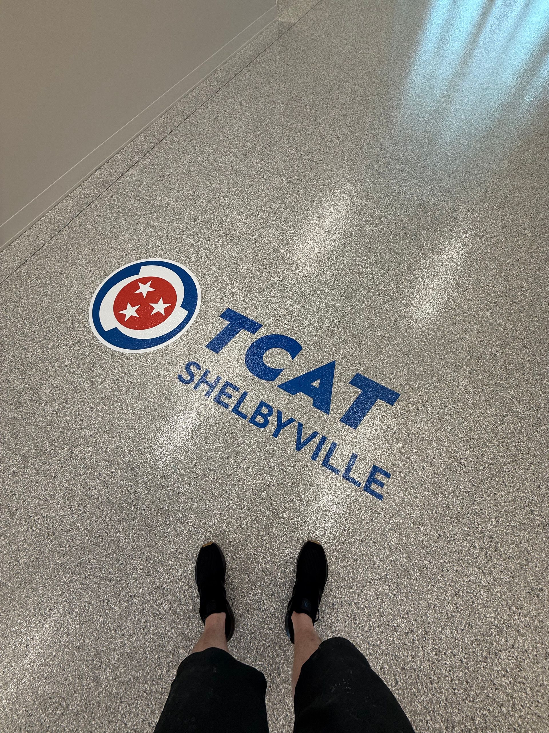 TAT Shelbyville logo on speckled floor, viewed from above with a person’s feet at bottom edge