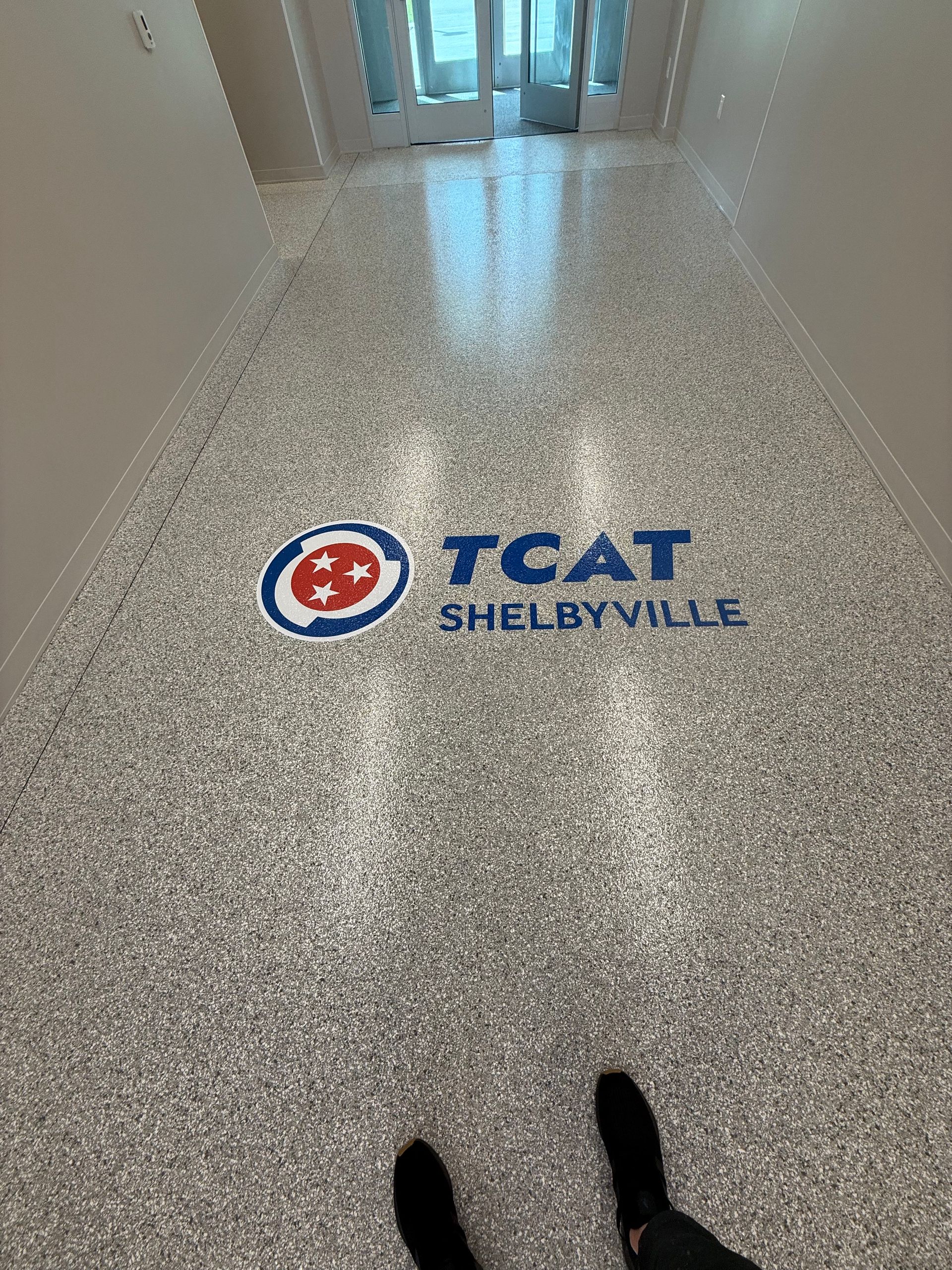 Hallway floor with TCAT Shelbyville logo and a person’s feet at the bottom edge