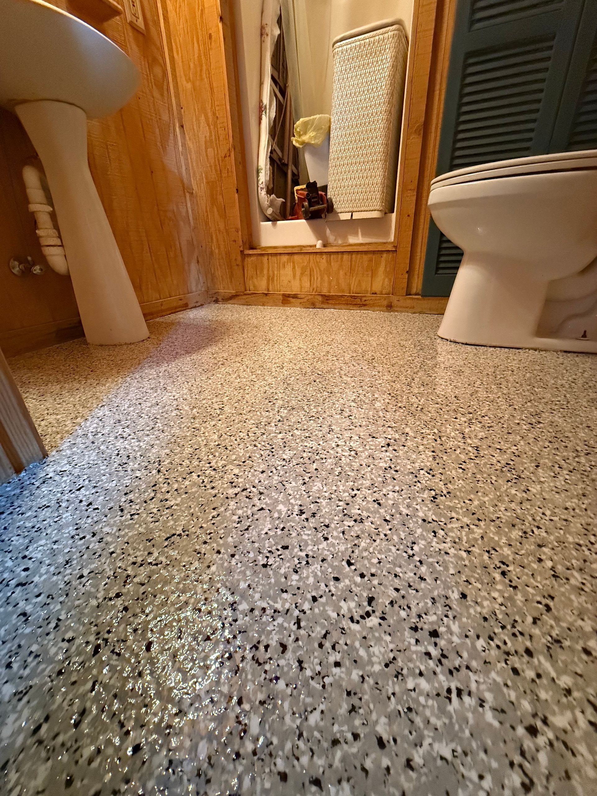 Small bathroom with speckled floor, toilet on each side, and mirror above a sink ahead.