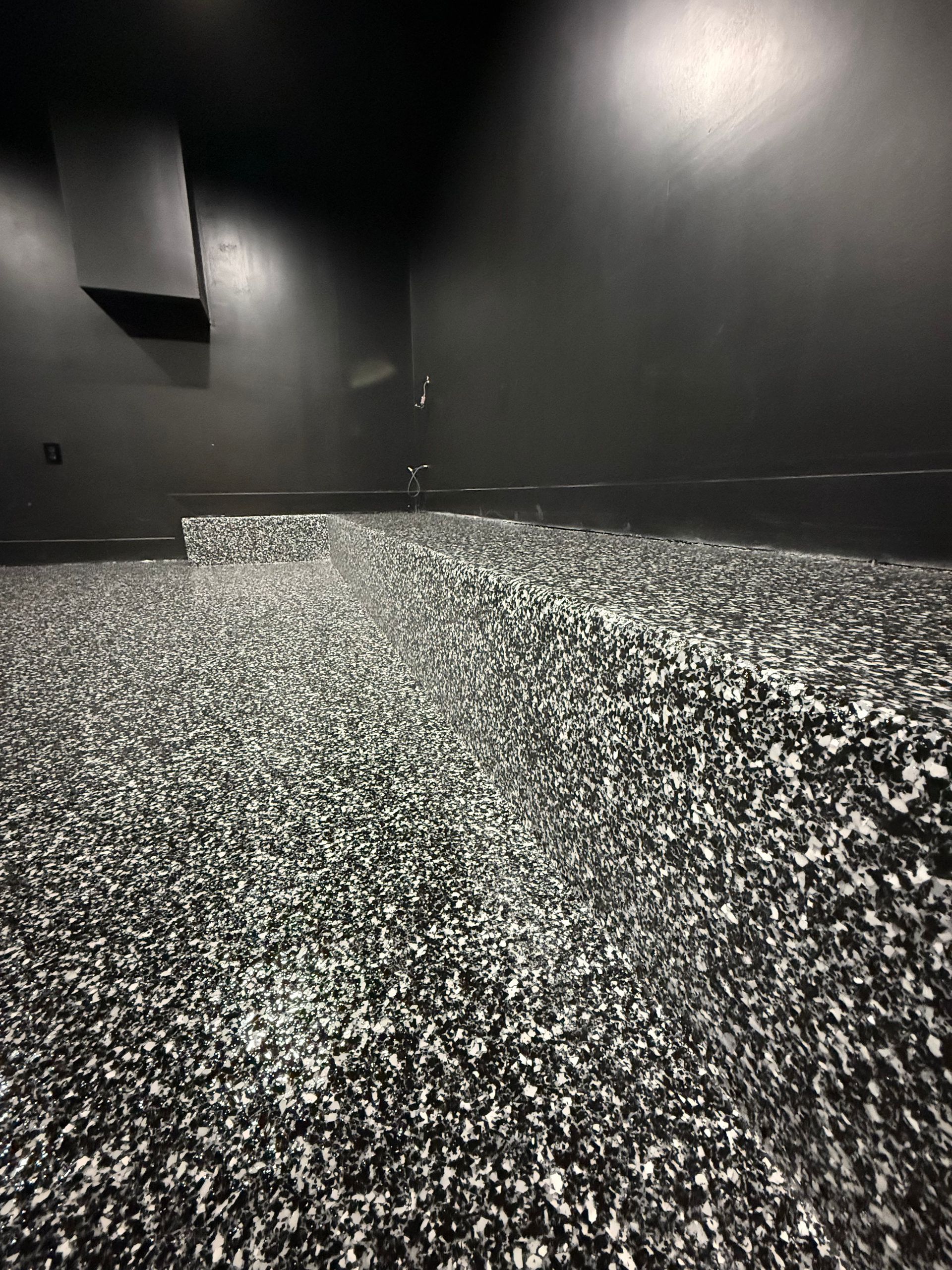 Empty dark room with black walls and a speckled black-and-white floor, lit by overhead spotlights